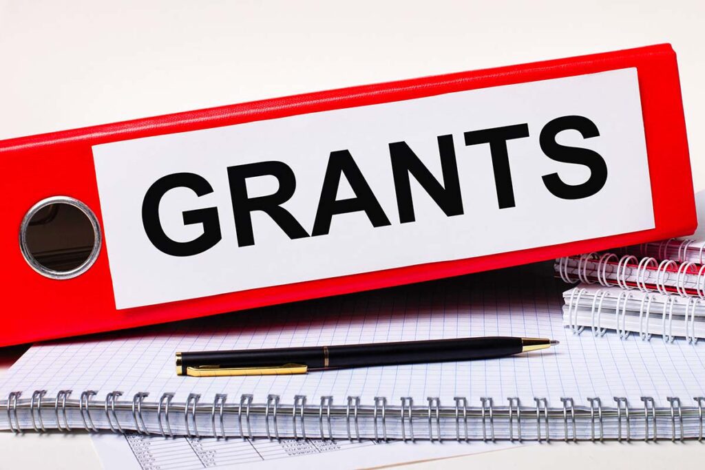 On the desktop are notebooks, a pen and a red folder for papers with the text GRANTS. Business concept